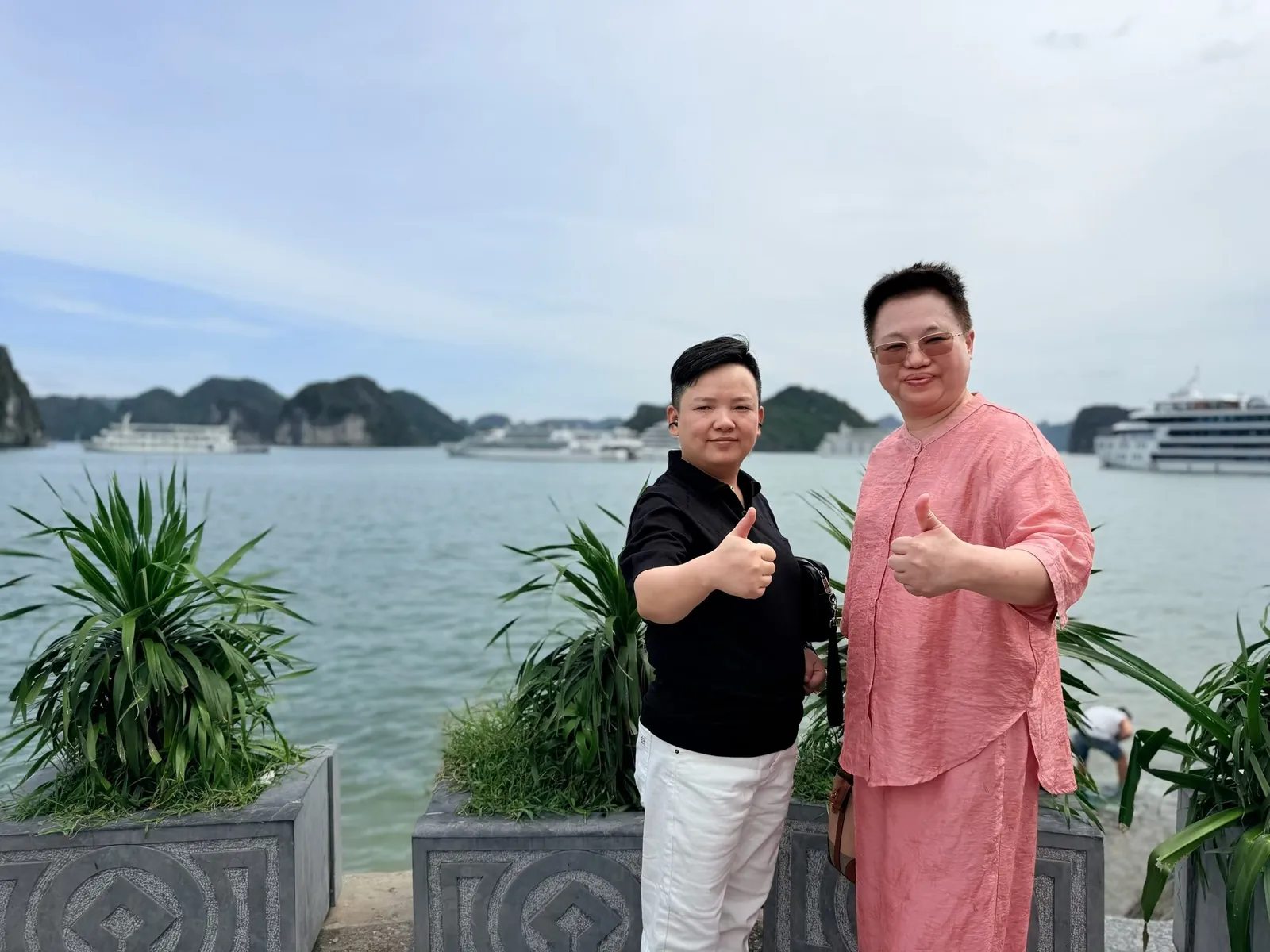 两人在下龙湾岸边竖起大拇指，身后是海面、游船和远处的群山