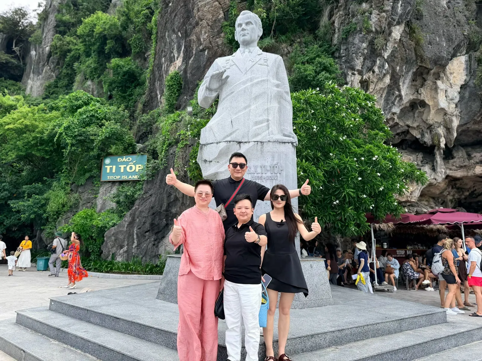 四人在 TITOP 岛季托夫雕像前竖起大拇指合影，背后是长满绿树的石灰岩山壁