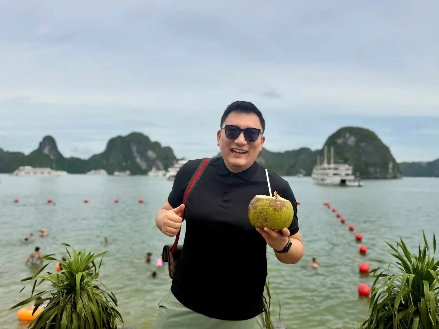 一名男子戴着墨镜手捧椰子,身后是下龙湾的海滩、游船和石灰岩岛屿